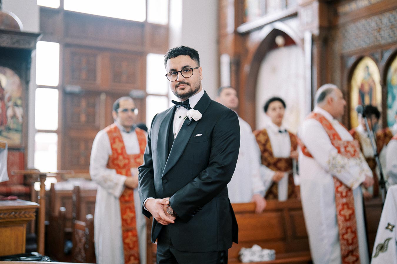 Man in a black tuxedo standing in a church with clergy in the background wearing ornate robes, highlighting a formal and ceremonial setting.