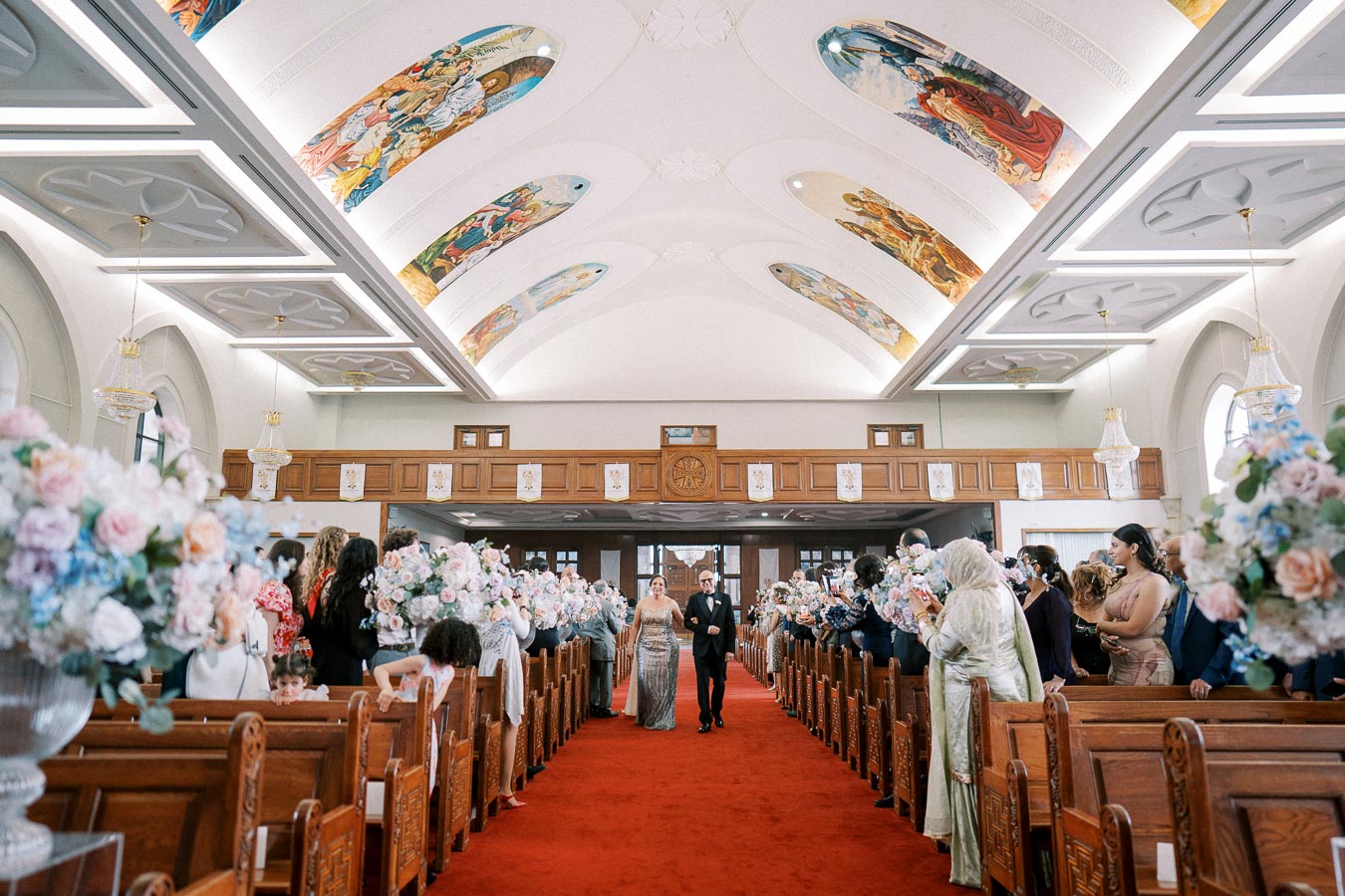 Wedding ceremony in an ornate church with decorative ceiling murals and floral arrangements, featuring a couple walking down a red-carpeted aisle surrounded by guests.