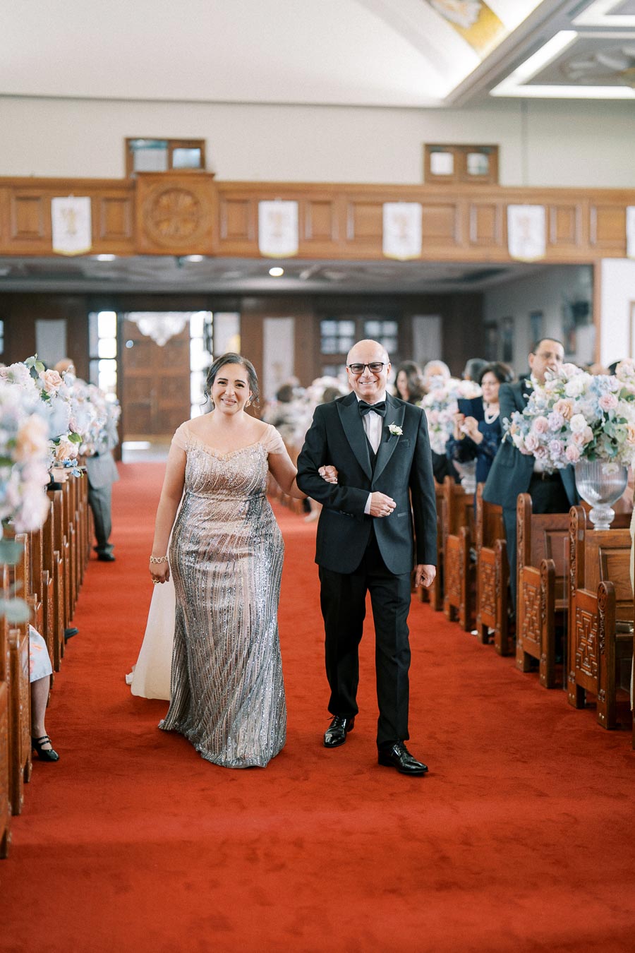 Wedding ceremony in a church with a woman in a sequined gown walking down the aisle, accompanied by a man in a tuxedo on a red carpet. Floral arrangements line the pews, creating an elegant atmosphere.