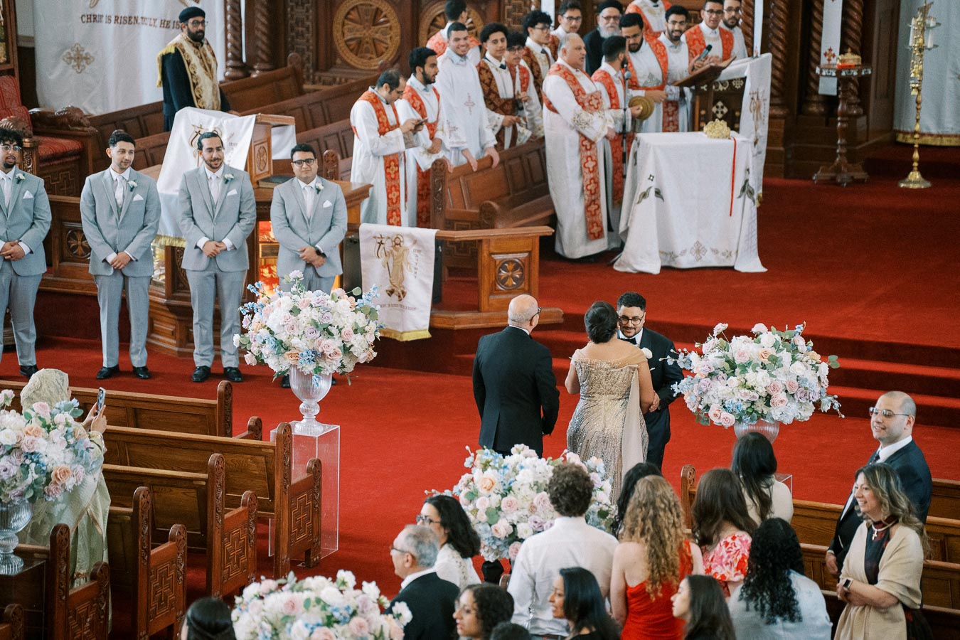 A traditional church wedding ceremony with guests seated inside, featuring a bridal couple greeting an elderly man at the altar. The scene includes clergy in ceremonial robes, groomsmen in light gray suits, and large floral arrangements on a red carpet.