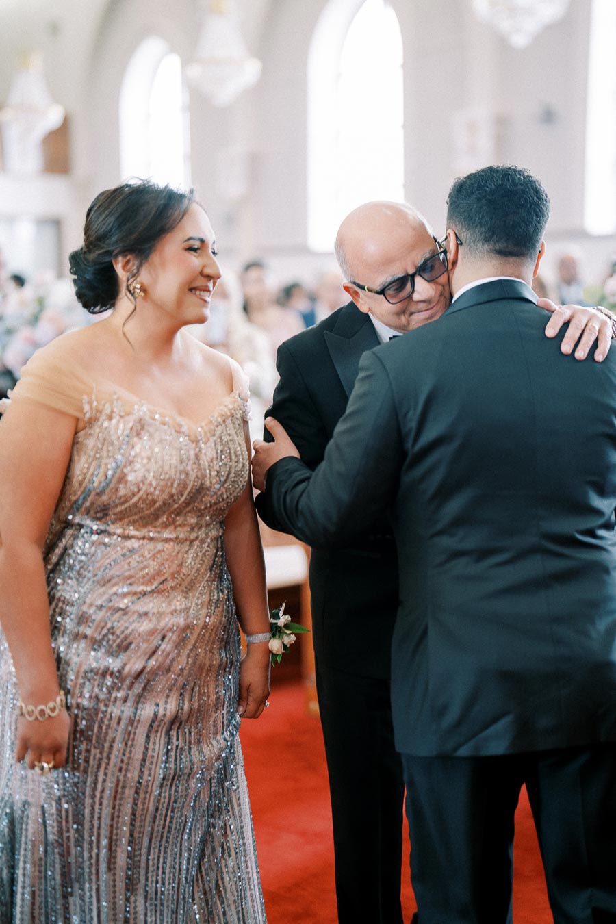 A woman in an elegant sequined gown smiles warmly as she stands beside two men wearing tuxedos, embracing in a softly lit, ornate venue.