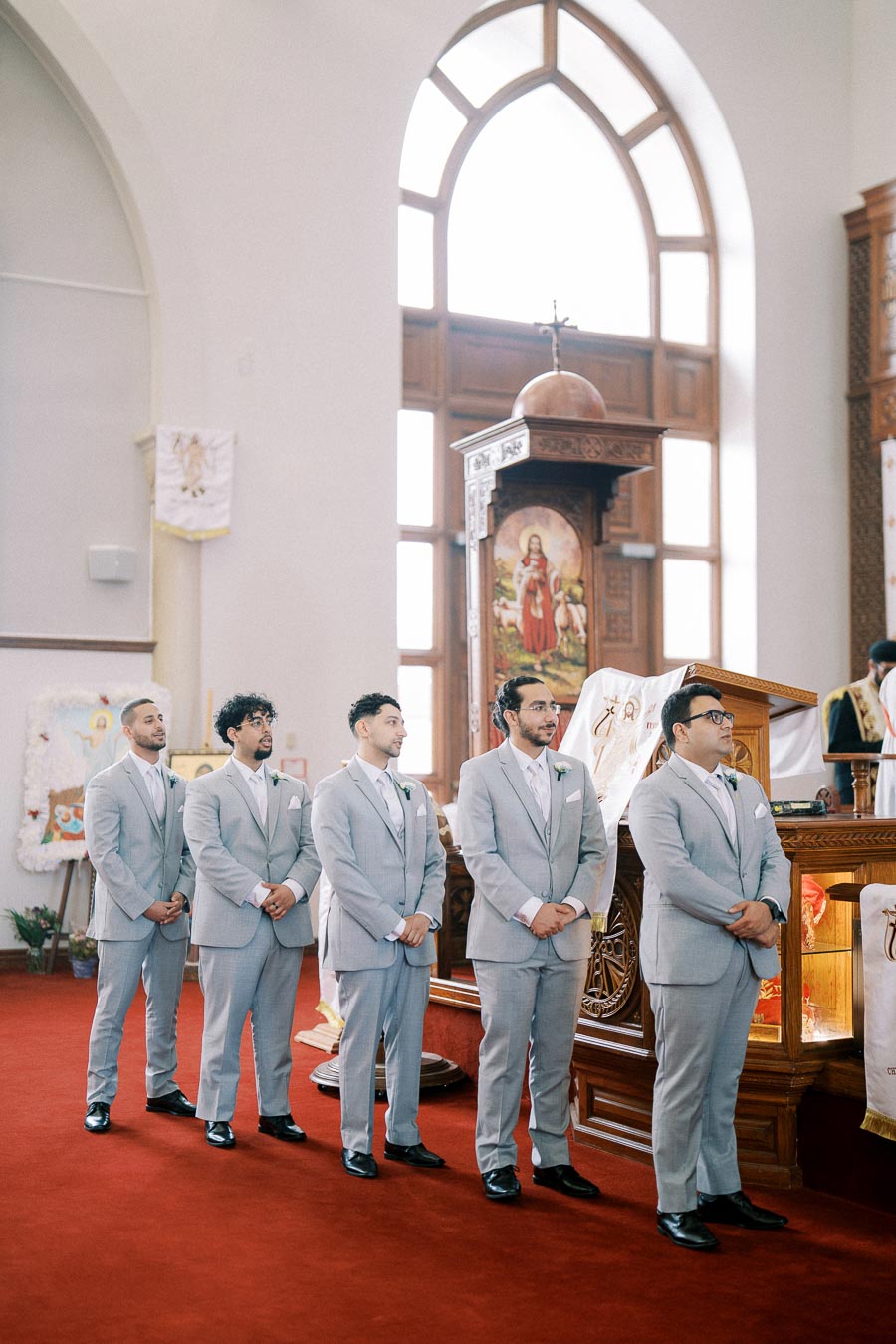 A group of groomsmen in matching gray suits stand in line inside a church, ready for a wedding ceremony.