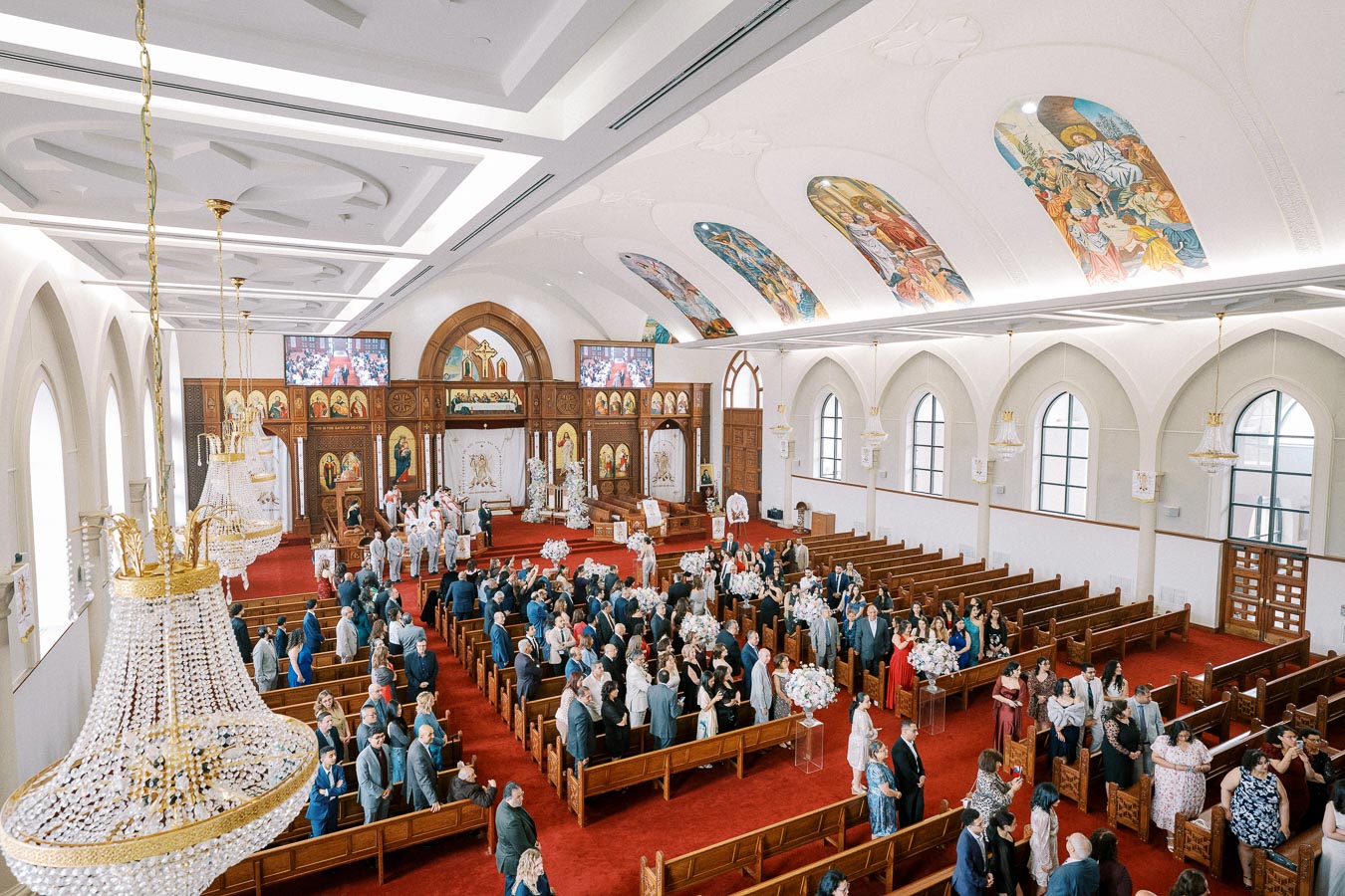 This image showcases a large, beautifully decorated church interior during a wedding ceremony. The spacious venue features ornate chandeliers, vibrant religious artwork on the ceiling, and rows of pews filled with elegantly dressed guests.