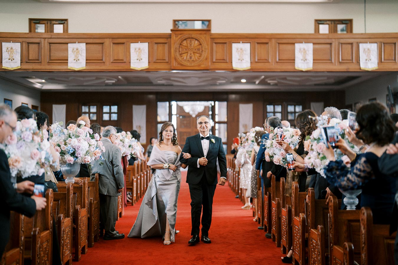 A couple walking down the aisle in a decorated church, surrounded by guests with flowers and smartphones, during a wedding ceremony.