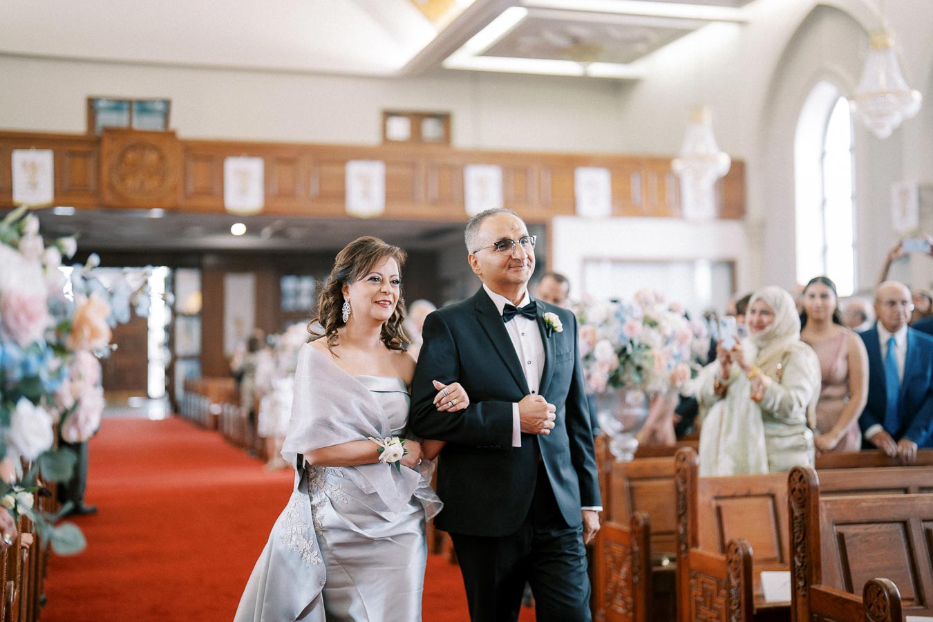 A couple elegantly dressed, walking down the aisle in a beautifully decorated church with floral arrangements and seated guests, during a formal ceremony.