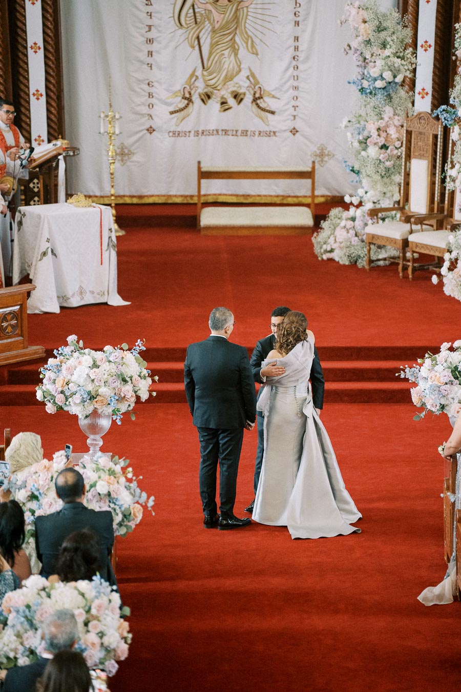 A wedding ceremony inside a church with a bride and groom embracing at the altar. The church features a red carpet, floral arrangements, and a religious tapestry with the words Christ is risen in the background.