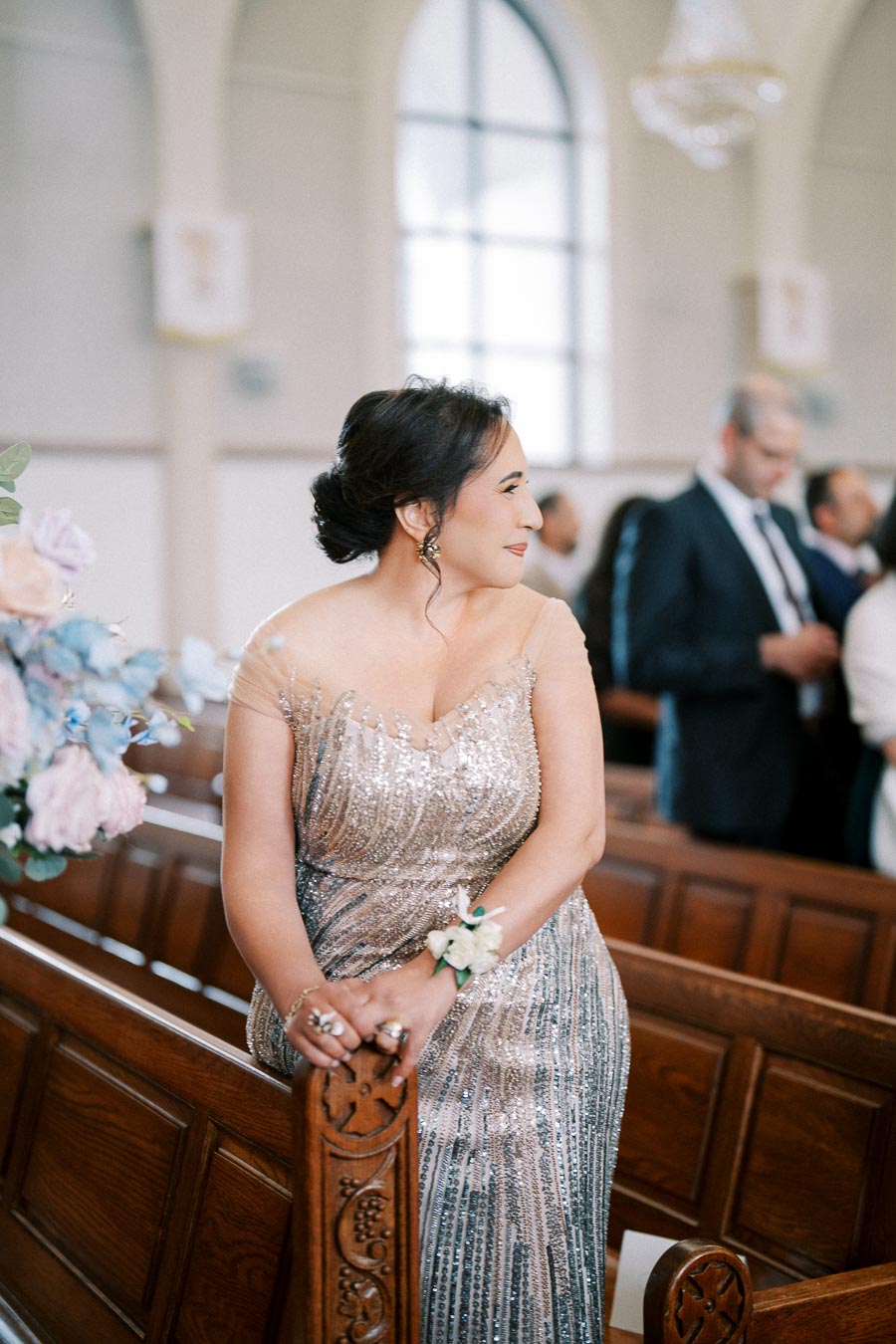 A woman in an elegant, shimmering silver gown rests against a wooden pew in a beautifully decorated church, adorned with pastel flowers, while guests converse in the background.