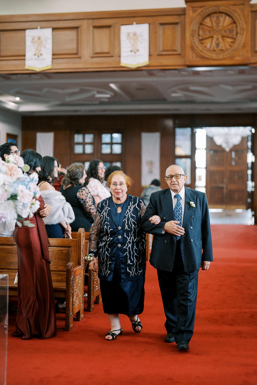 Elderly couple walking down the aisle at a wedding ceremony, surrounded by seated guests in a beautifully decorated church with wooden accents and red carpet.