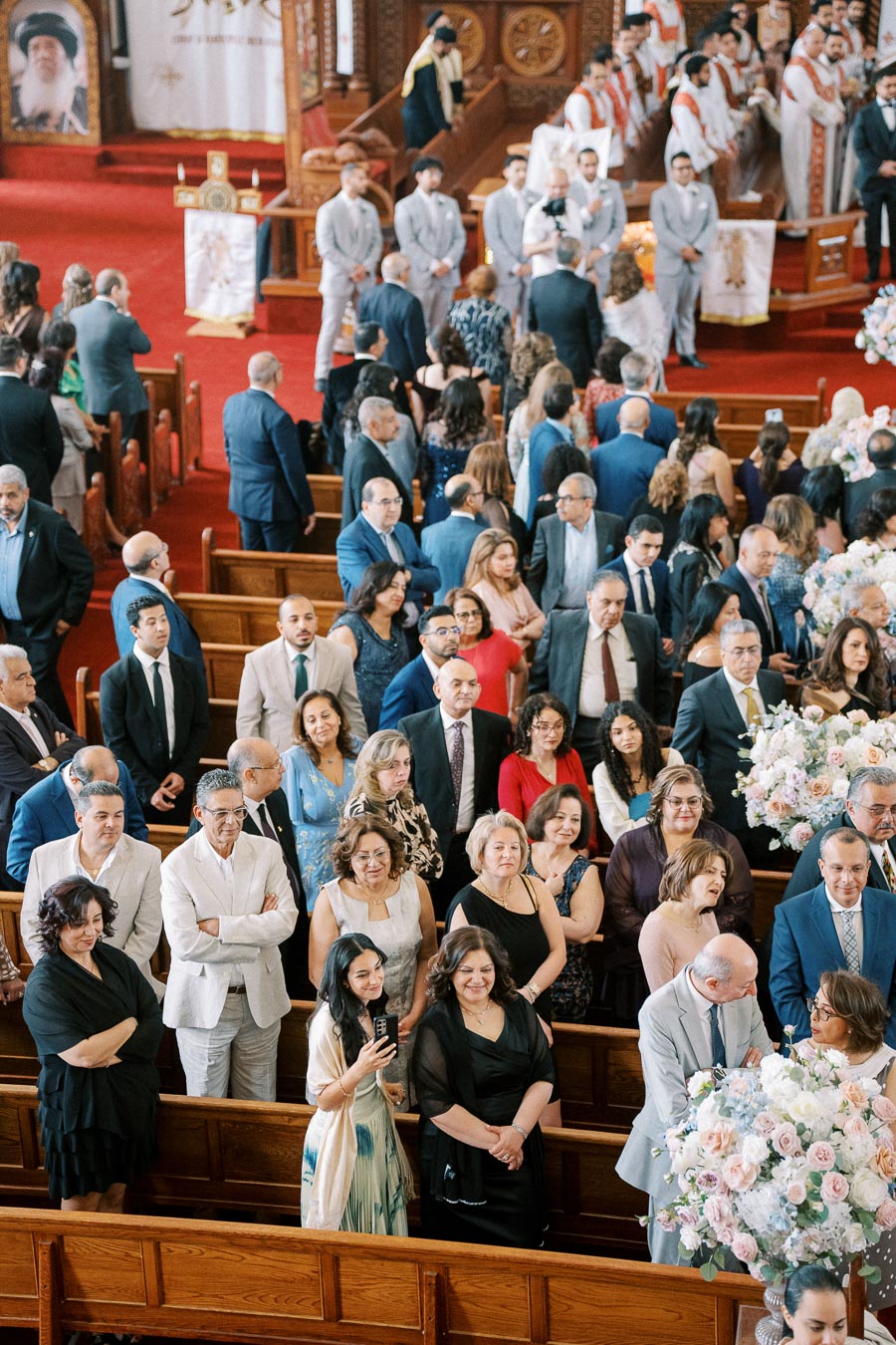 A congregation gathers in an ornate church with red carpeted aisles, as people dressed in formal attire participate in a ceremony. Floral arrangements and religious figures are visible at the front.