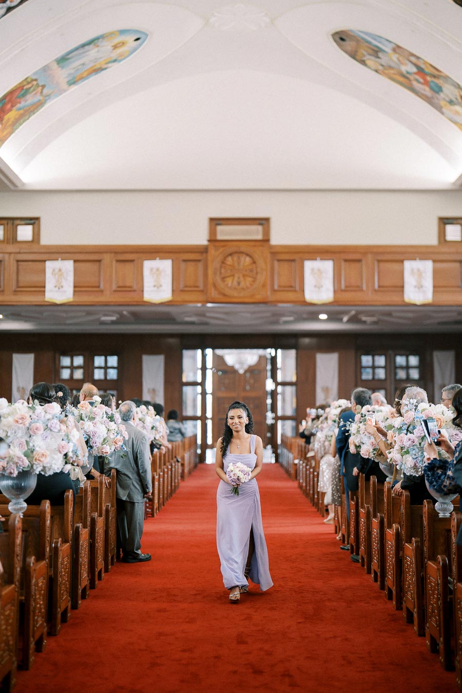 Bridesmaid walking down the aisle in a church, wearing a lavender dress and holding a bouquet, with guests seated on both sides and floral decorations along the pews.