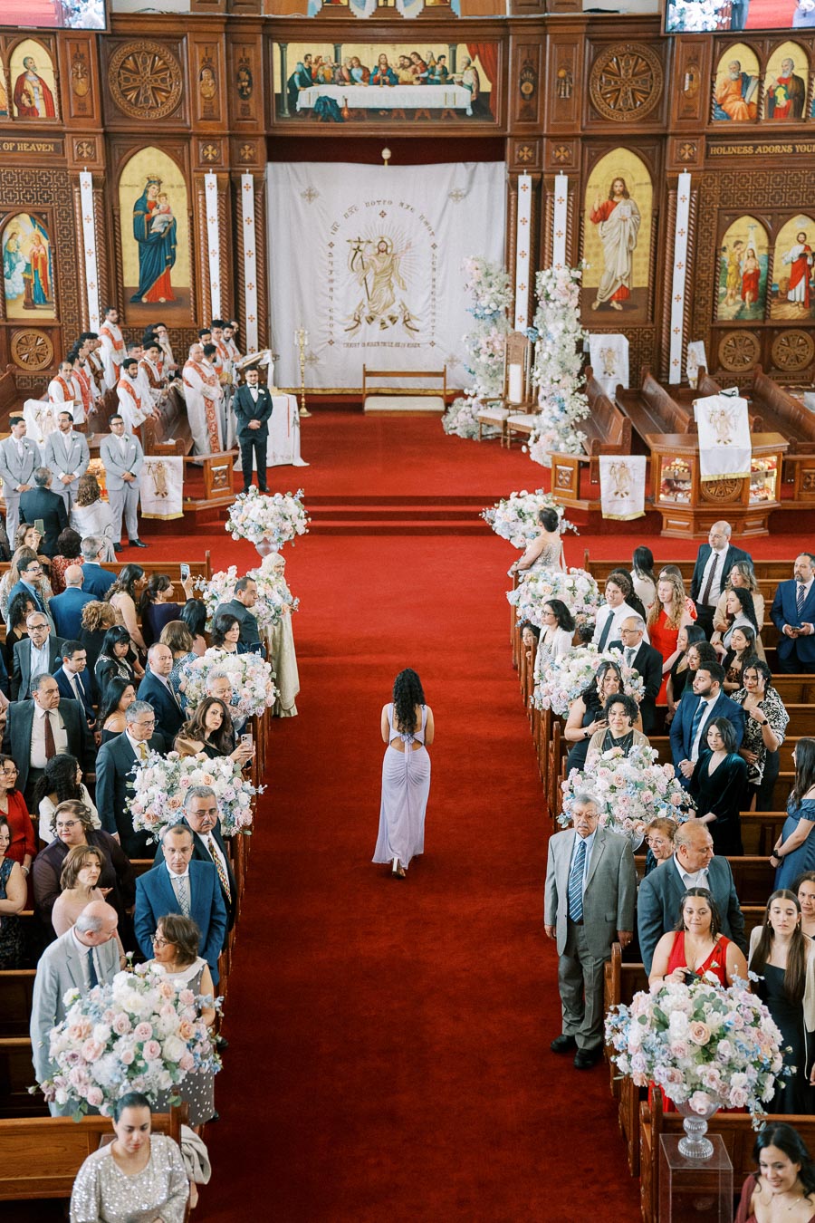 A grand wedding ceremony takes place in an ornately decorated church with a red carpet aisle, surrounded by guests in formal attire and floral arrangements, highlighting the church’s detailed wooden architecture and religious artwork.