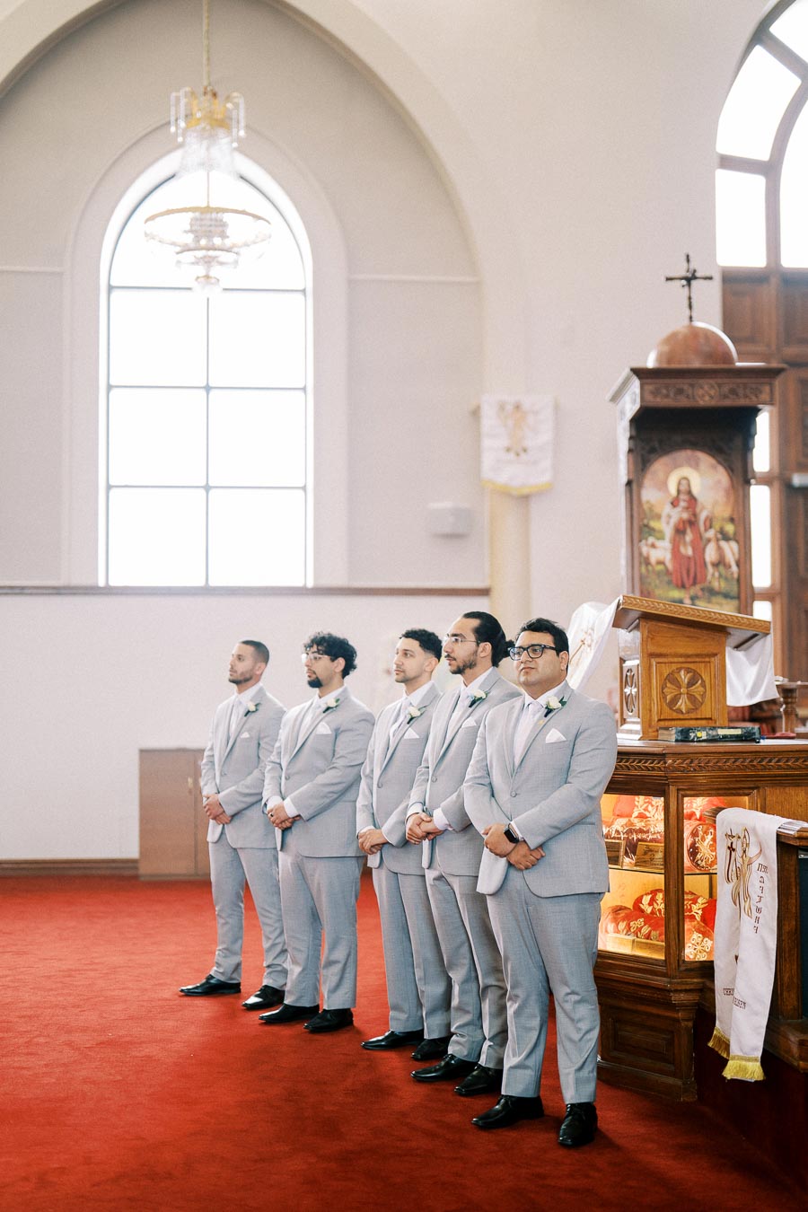 Group of groomsmen in light gray suits standing in a church setting, red carpet and stained glass window in the background, next to a decorated wooden pulpit.
