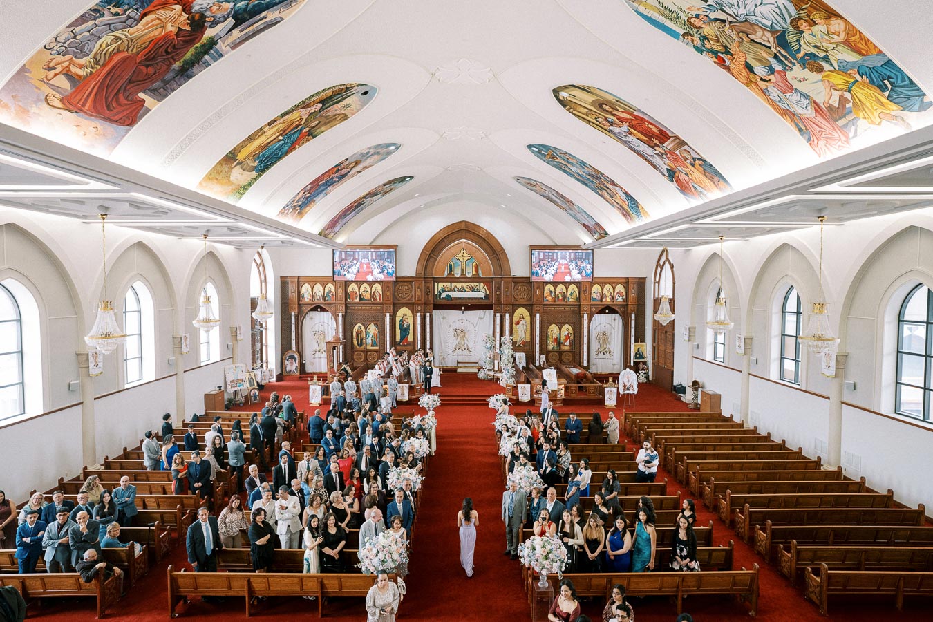 Wide-angle view of a beautifully decorated church interior during a wedding ceremony, featuring colorful ceiling murals, ornate chandeliers, and guests seated in wooden pews on either side of a red carpet aisle.