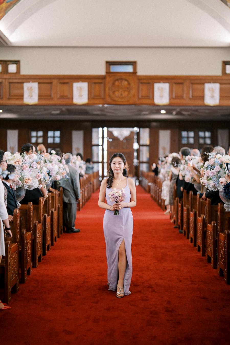 Bridesmaid in purple gown walking down the aisle of a beautifully decorated church, holding a bouquet of pastel flowers, during a wedding ceremony.