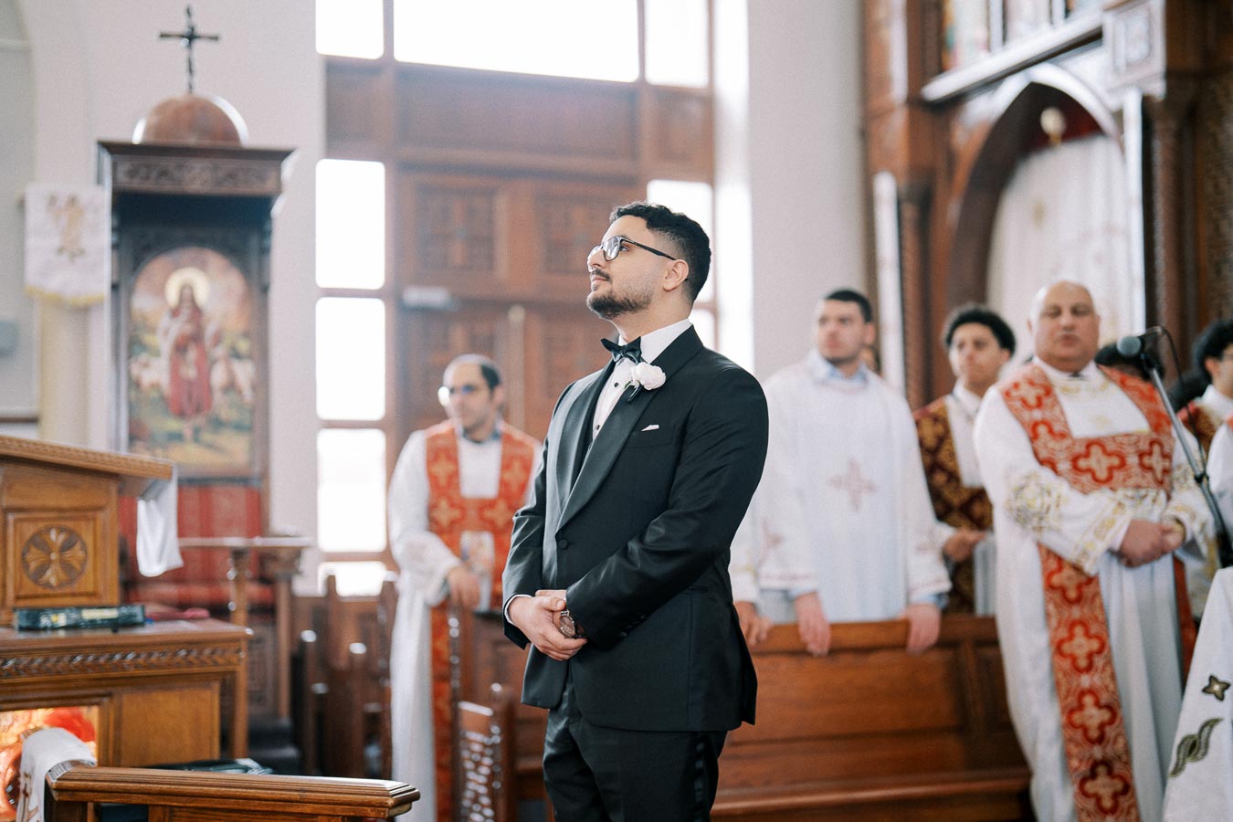 Man in formal suit standing inside a church alongside clergy in traditional robes, with wooden pews and religious decor in the background.