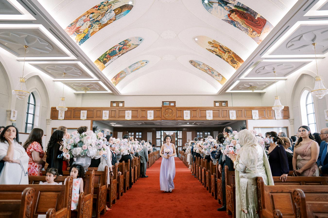 Elegant church wedding ceremony with a purple-dressed bridesmaid walking down a flower-adorned aisle, surrounded by guests seated on wooden pews beneath a beautifully decorated ceiling.