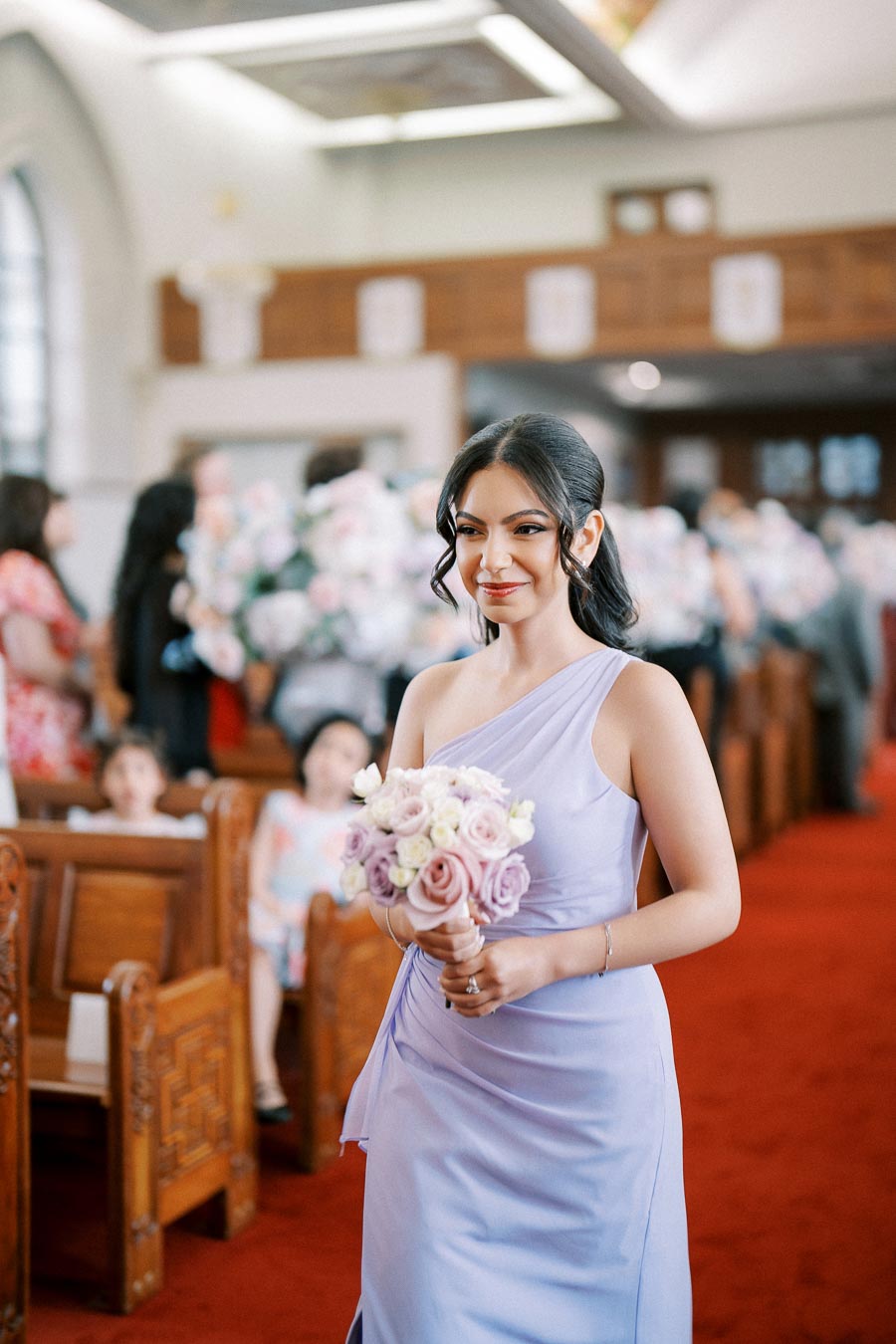 A woman in a light purple dress walks down a red-carpeted aisle holding a bouquet of pastel roses at a formal event inside a decorated venue with wooden seating and floral arrangements in the background.