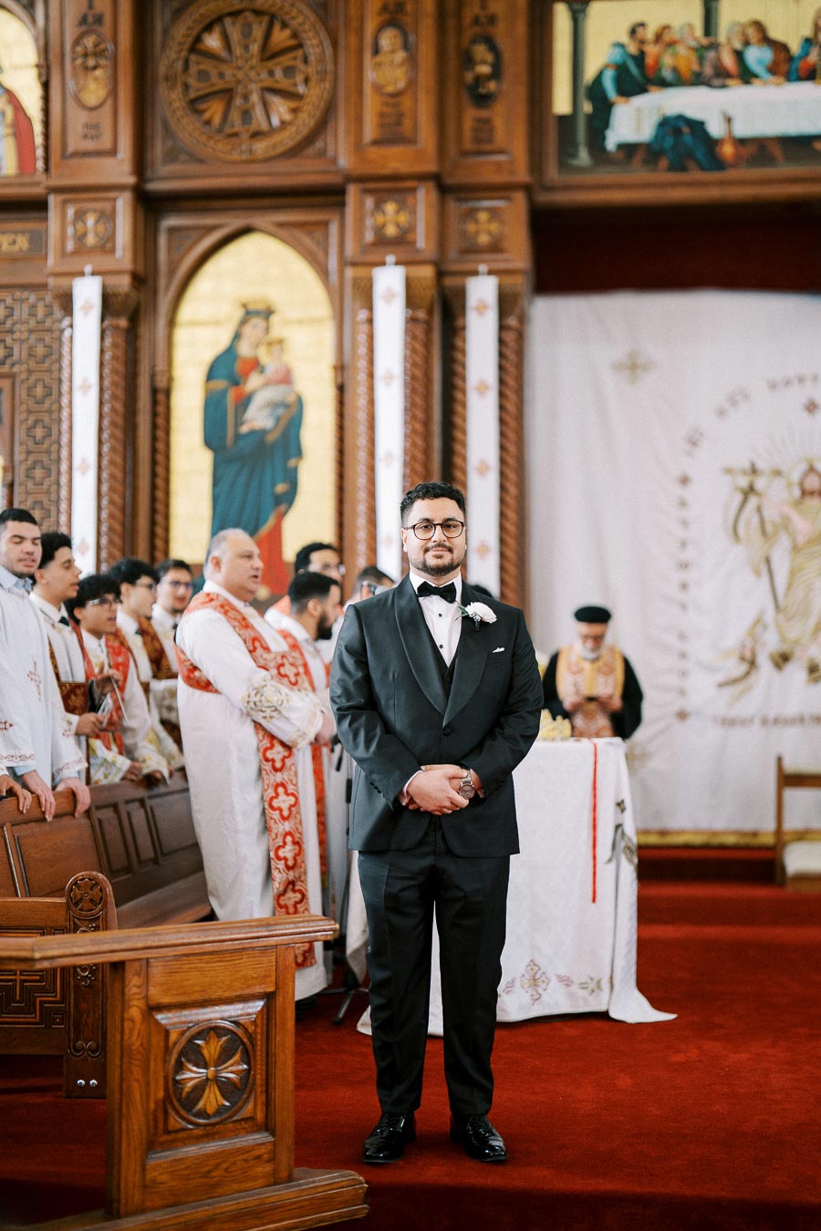 A man in a black tuxedo stands inside a beautifully decorated church with red carpeting and ornate wooden furnishings, surrounded by clergy in ceremonial robes during a religious event.