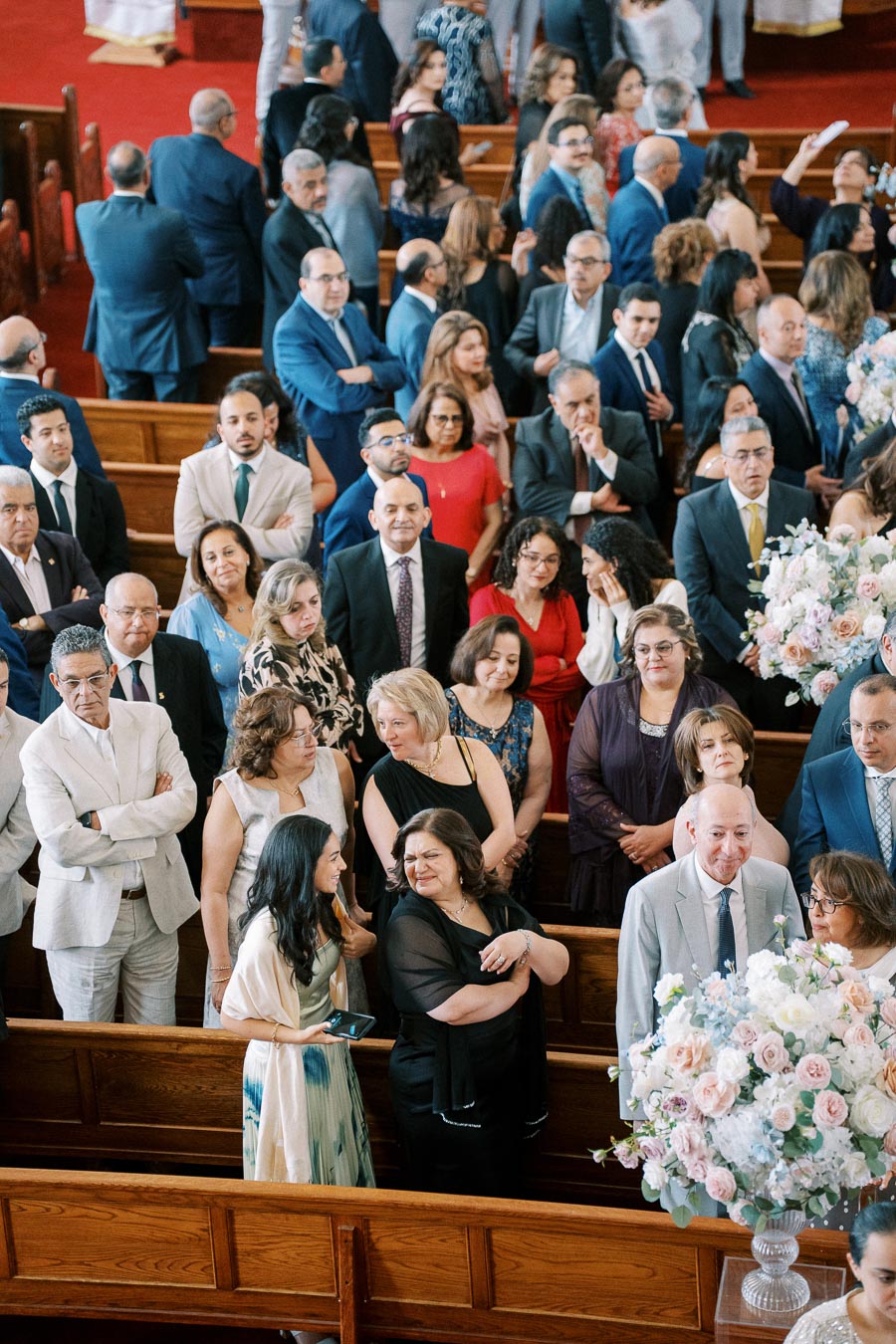 A large group of well-dressed people gather inside a church, engaging in conversation and waiting for an event to begin. The church pews and floral arrangements create an elegant setting, suggesting a formal ceremony or celebration.