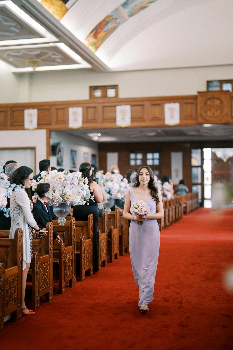 Bridesmaid in a lavender dress walking down the aisle holding a bouquet of pink and white flowers in a beautifully decorated church with wooden pews and floral arrangements.