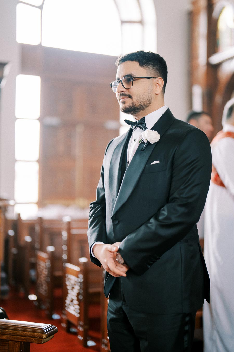 A groom in a black tuxedo with a white boutonniere stands in a sunlit church, waiting expectantly at the altar.
