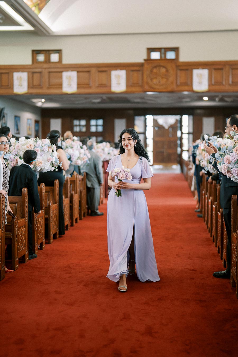 Bridesmaid walking down the aisle in a lavender dress, holding a bouquet of purple and white flowers, during a church wedding ceremony.
