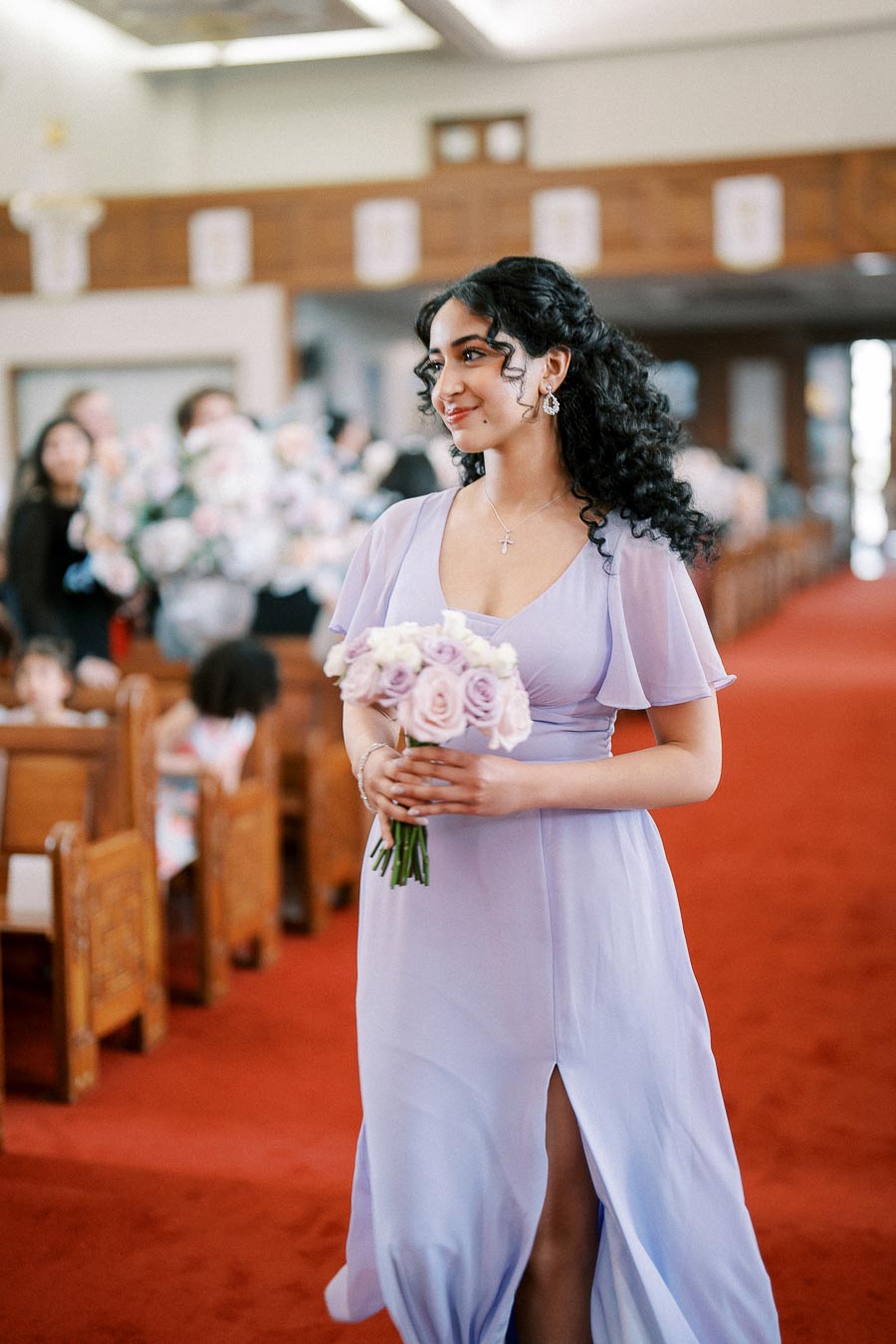 Young woman in a lavender bridesmaid dress holding a bouquet of roses, smiling while walking down the aisle at a wedding ceremony in a church.