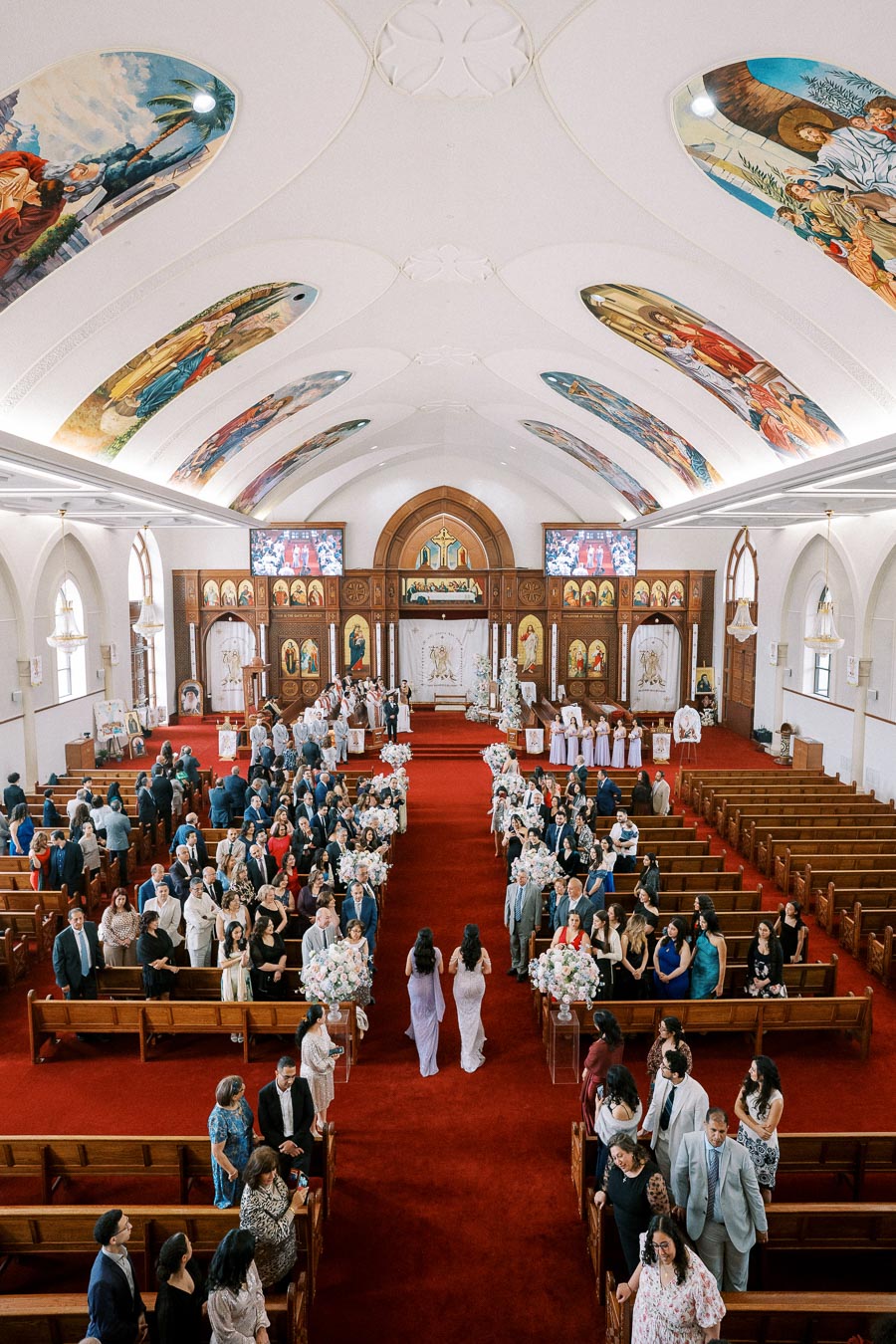 Interior of a beautifully decorated church filled with wedding guests, featuring ornate ceiling murals, a red carpet aisle, and elegant floral arrangements lining the pews.