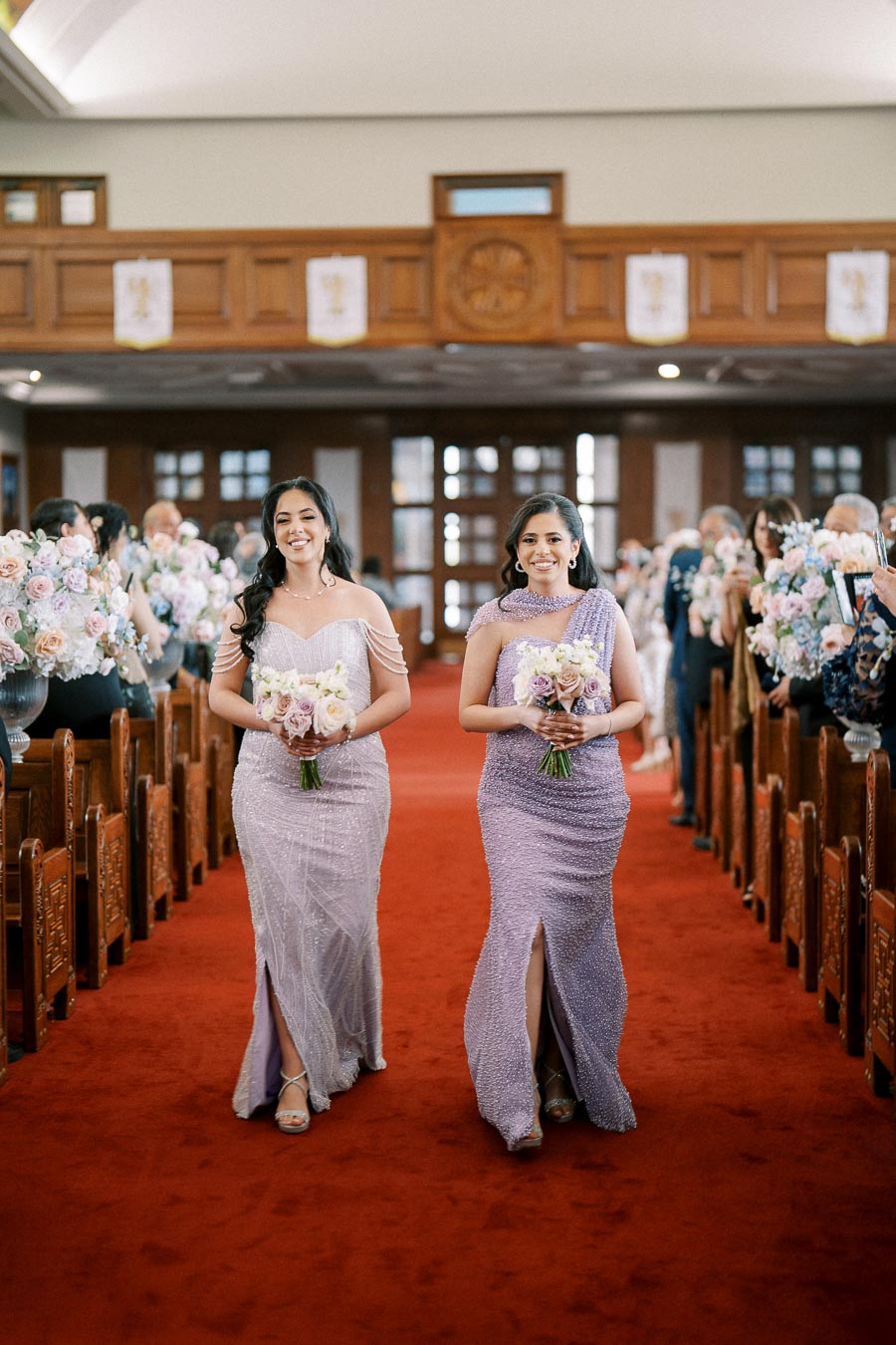 Two bridesmaids in matching elegant lavender gowns walk down a red carpeted aisle, holding bouquets, with a wedding ceremony in progress in the background.