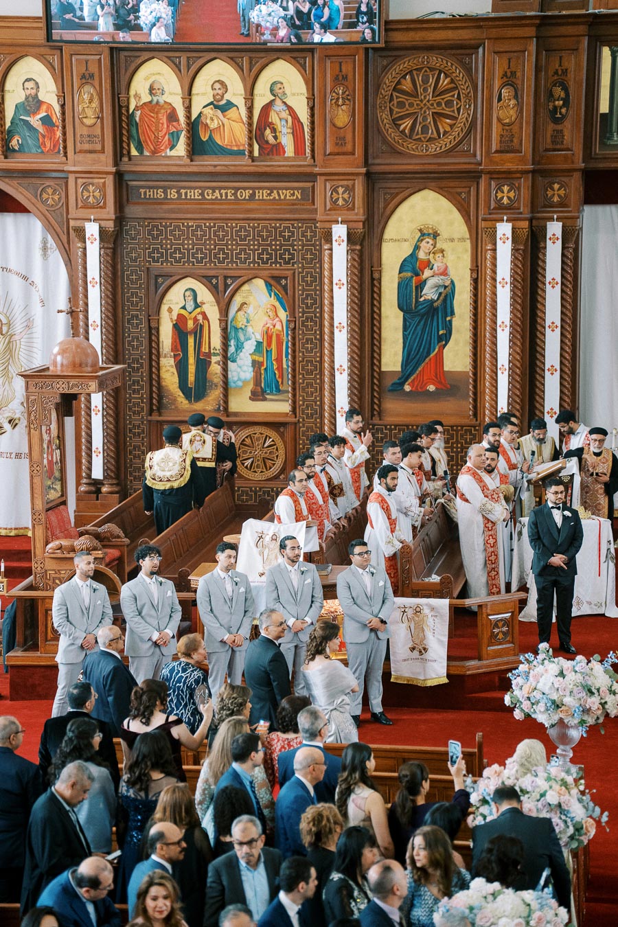 A vibrant and ornate church ceremony featuring priests in white and red robes, groomsmen in light gray suits, and guests gathered in celebration. The setting is adorned with religious icons and floral arrangements, with intricate woodwork and artworks creating a reverent atmosphere.