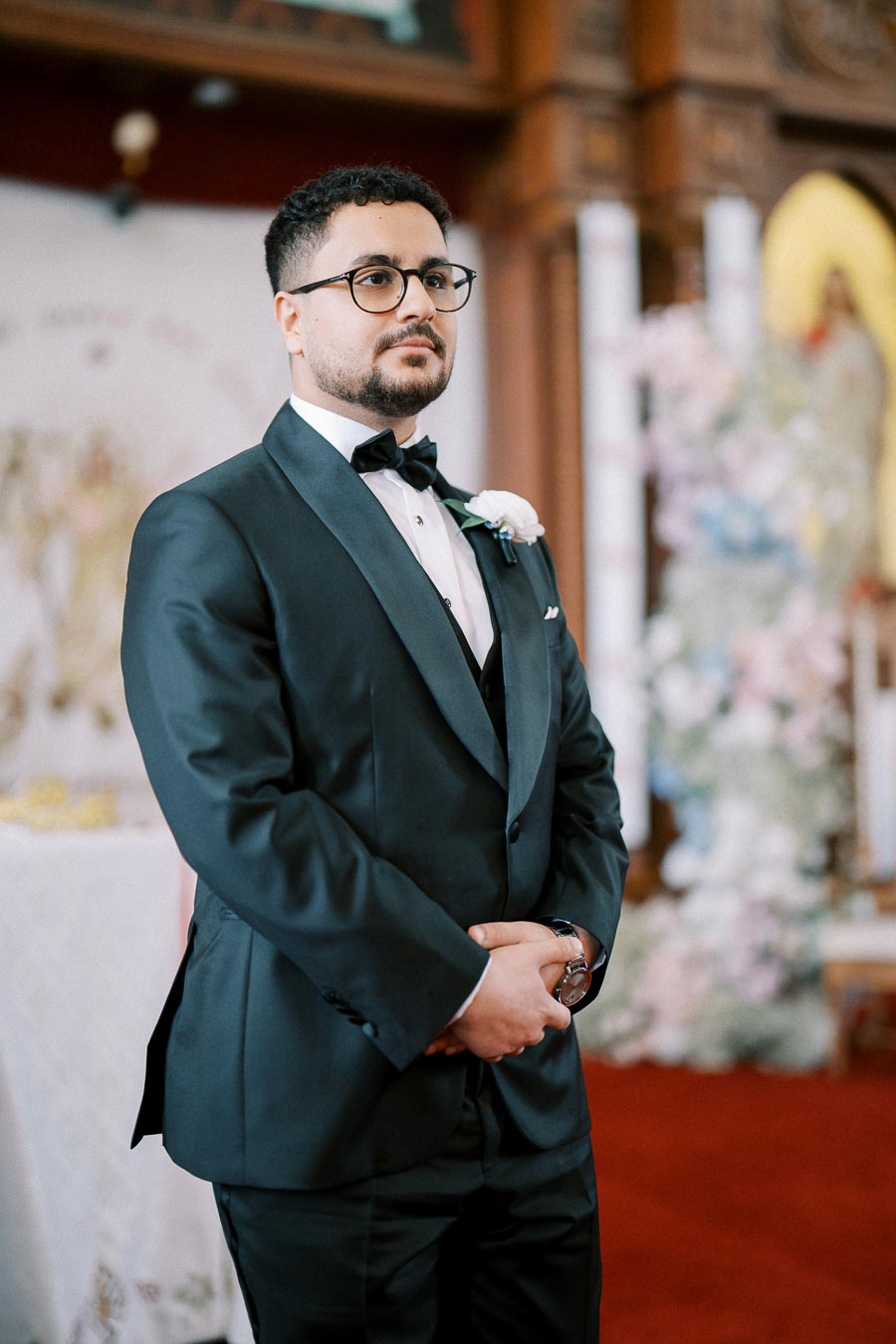 A groom in a black tuxedo and bow tie stands in a church, looking composed and ready for the wedding ceremony.