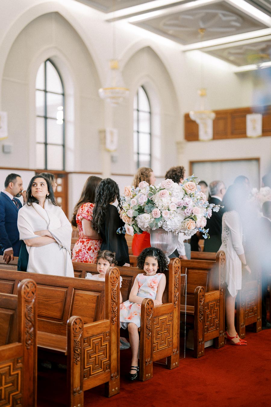 A group of people, including children, seated in ornate wooden pews in a brightly lit, large hall with arched windows, featuring a large floral arrangement.