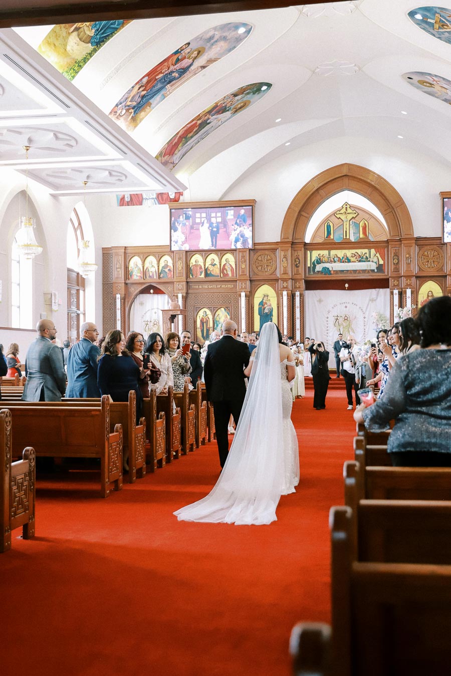A bride walks down the aisle with her father in a beautifully decorated church, adorned with religious artwork and a vibrant red carpet, while guests look on during a wedding ceremony.