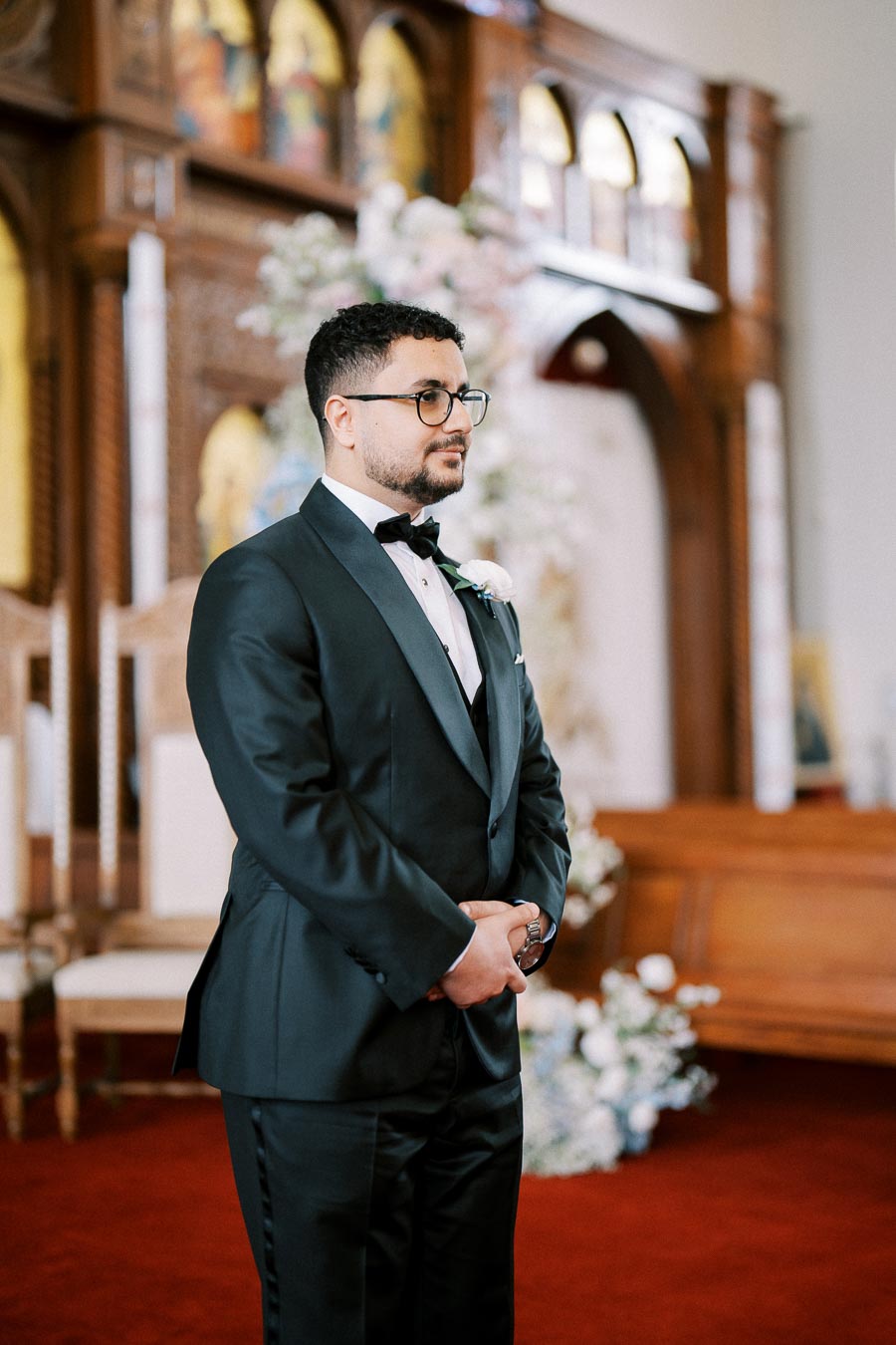 A groom in a sleek black tuxedo stands in a beautifully decorated church, surrounded by elegant floral arrangements and rich wooden architecture, eagerly awaiting his wedding ceremony.