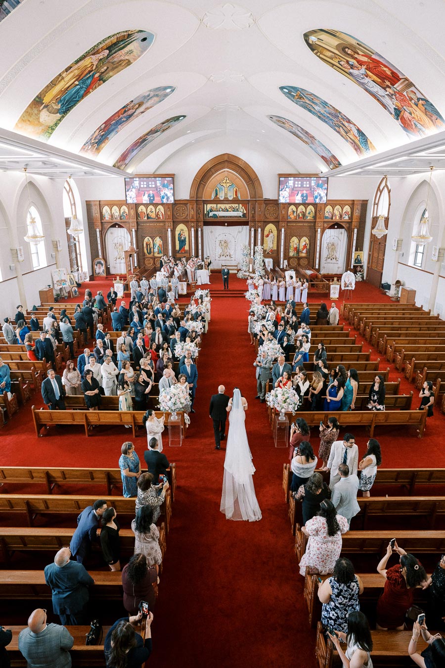 A bride walking down the aisle in a beautifully decorated church with a vibrant red carpet, surrounded by seated guests and floral arrangements, under ornate painted ceilings.