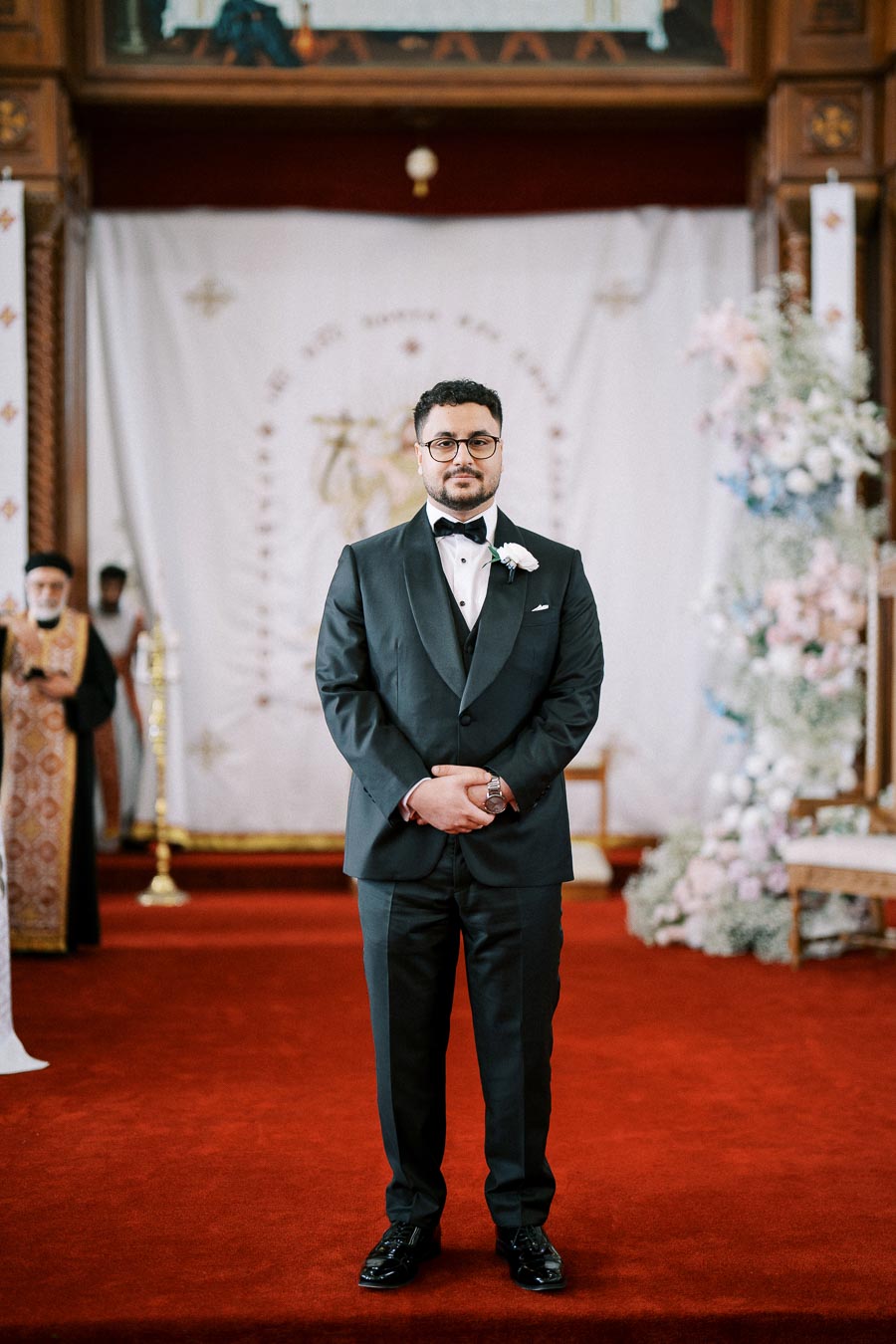 A groom in a black tuxedo standing on a red carpet in an elegant wedding venue with floral decorations and a white backdrop in the background.