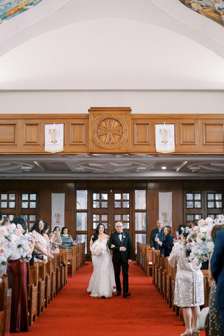 A bride in a white wedding gown walks down the red-carpeted aisle of a church, arm-in-arm with an older man in a black tuxedo. Guests stand on either side capturing the moment with their phones. The church interior features wooden pews and decorative wall panels.