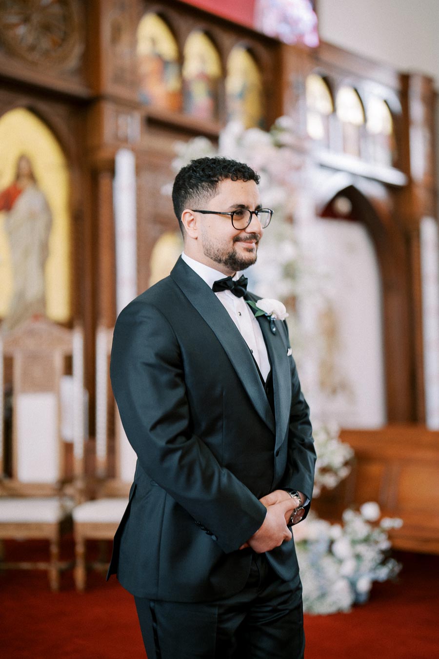 Groom in a black tuxedo with bow tie standing in a church setting, smiling and waiting at the altar; intricate wooden architecture and floral arrangements in the background.