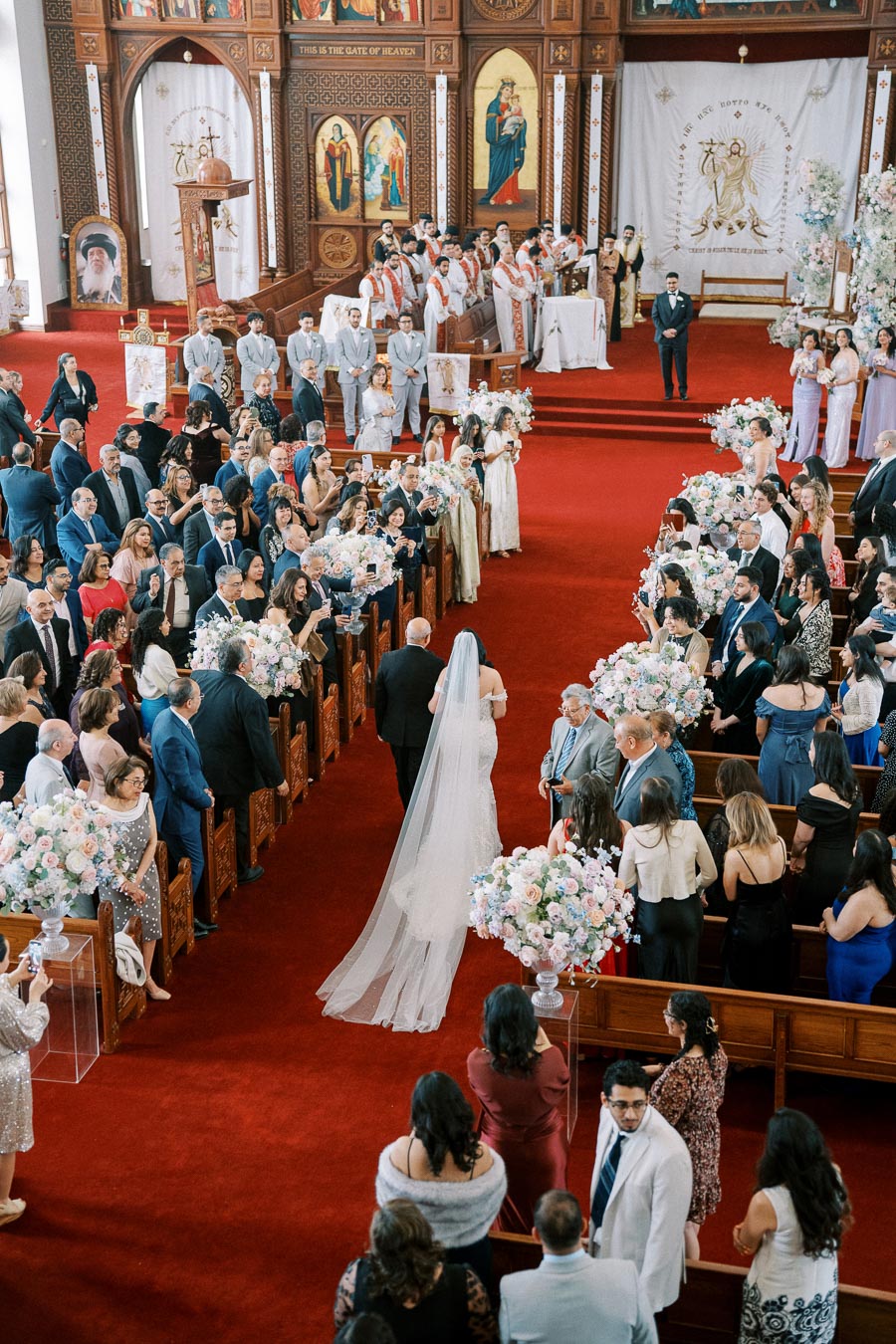 A bride with a long veil walking down the aisle of a beautifully decorated church, filled with guests dressed in formal attire, colorful floral arrangements, and a religious ceremony taking place at the altar.