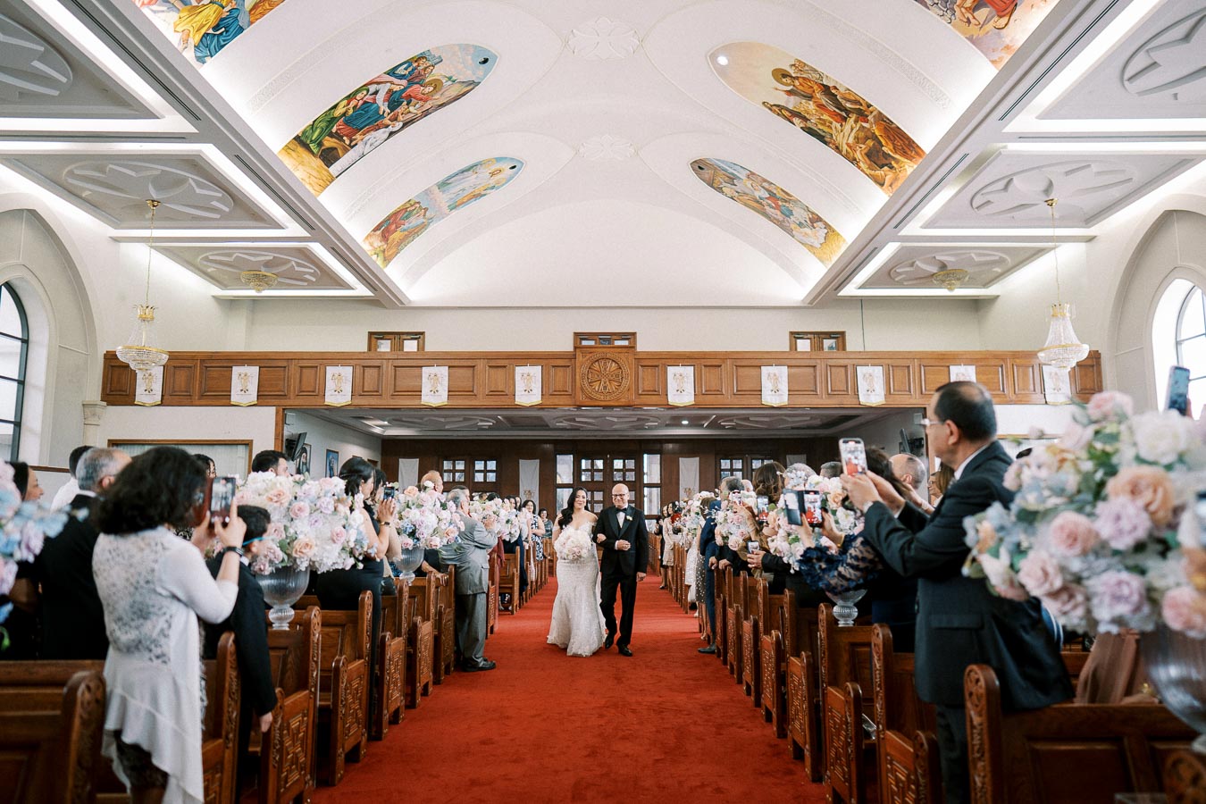 Bride walking down the aisle with her father in a beautifully decorated church, featuring ornate wooden pews and elegant floral arrangements. Guests capture the moment with their phones, while vibrant ceiling murals add a touch of color to the ceremony.