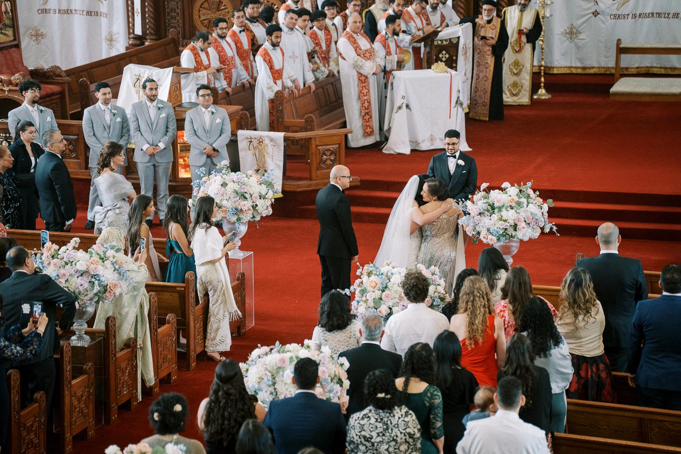Elegant church wedding ceremony with bride and groom, surrounded by family, friends, and officiants, in a richly adorned setting featuring red carpets and floral displays.