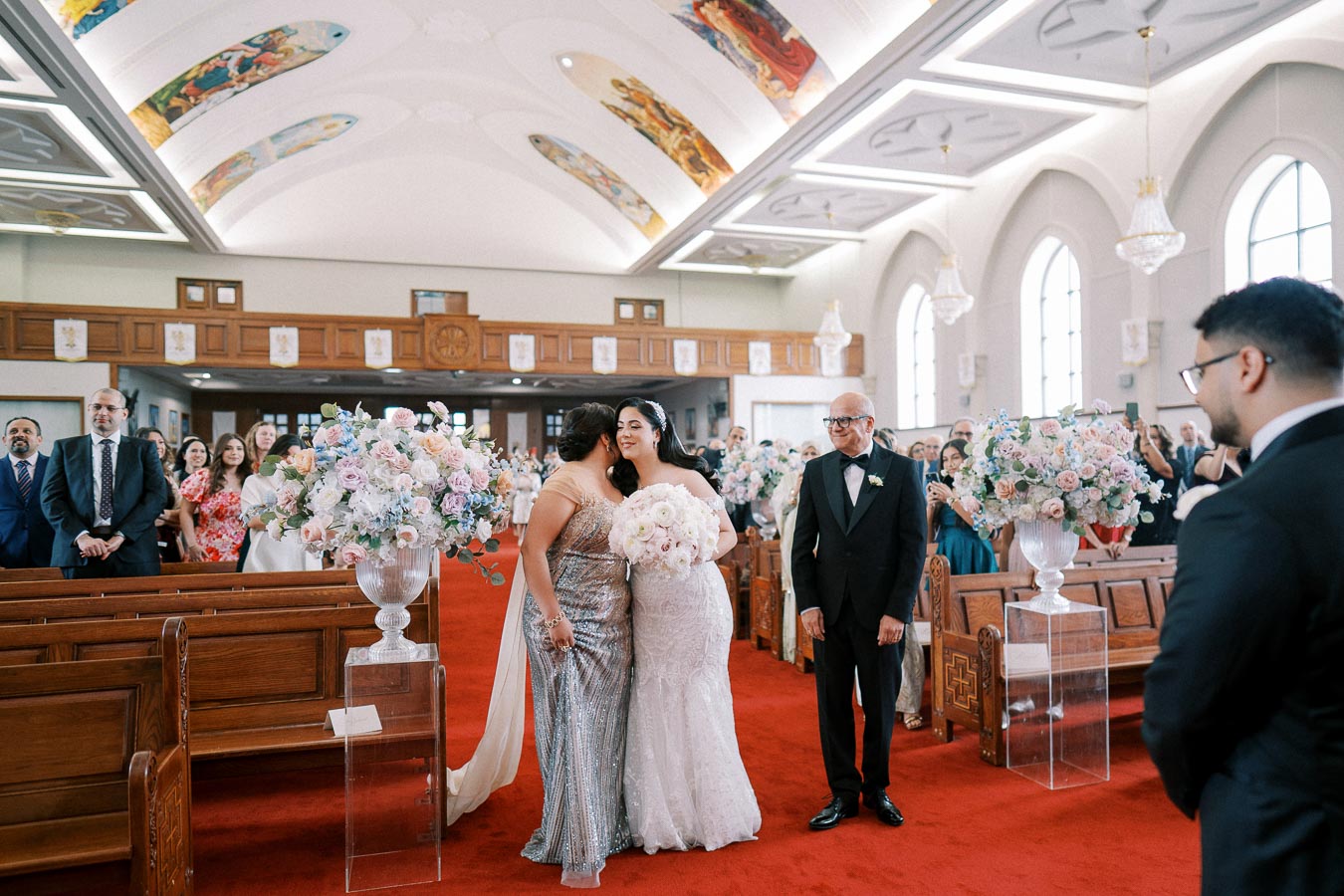 A bride in a white wedding gown embraces a woman in a sparkling dress inside a beautifully decorated church, adorned with floral arrangements and attended by family and friends standing in the pews.