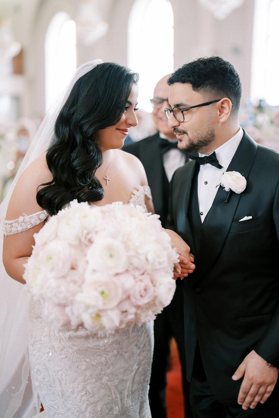 A bride and groom share a joyful moment during their wedding ceremony, with the bride holding a large bouquet of pale pink and white flowers, both dressed in elegant attire.
