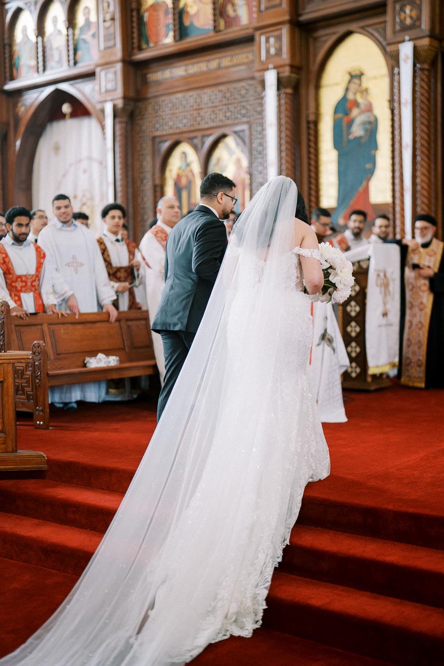 A bride and groom walk up the aisle in a beautifully adorned church, surrounded by clergy in ceremonial robes, with stained glass and intricate woodwork in the background.