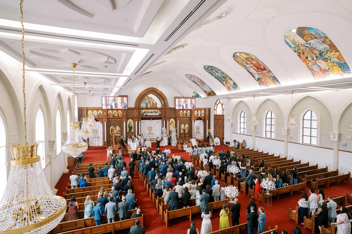 Interior view of a large, ornate church during a wedding ceremony, featuring a congregation seated in wooden pews, vibrant religious artwork on the ceiling, and elaborate chandeliers, emphasizing the ceremony's solemnity and elegance.