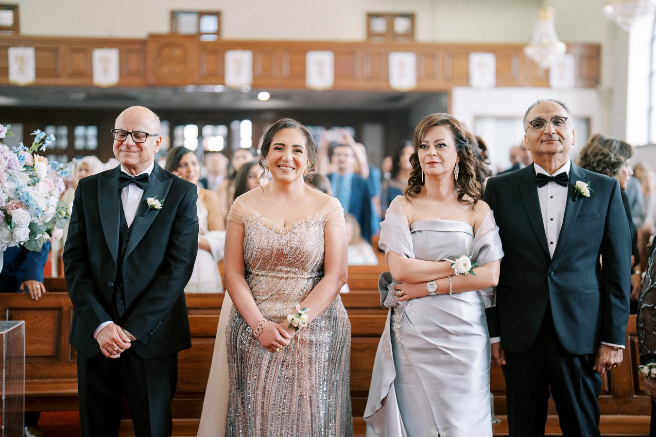 A group of elegantly dressed people smiling during a formal event inside a church, with flower arrangements and a congregation in the background.