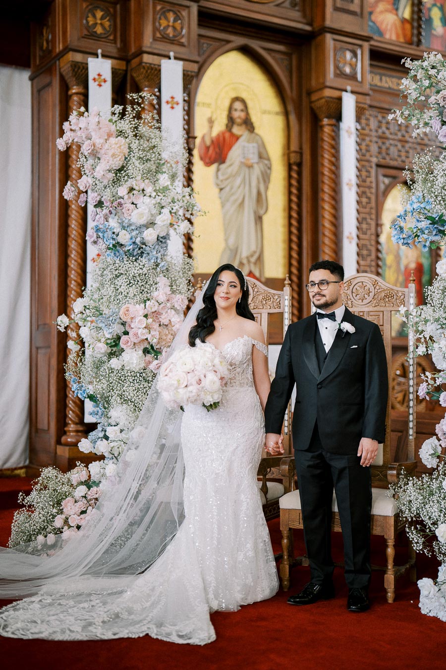 A bride in a white lace gown and veil holds a bouquet of flowers, standing beside a groom in a black tuxedo, in a richly decorated church setting with floral arrangements and religious iconography in the background.