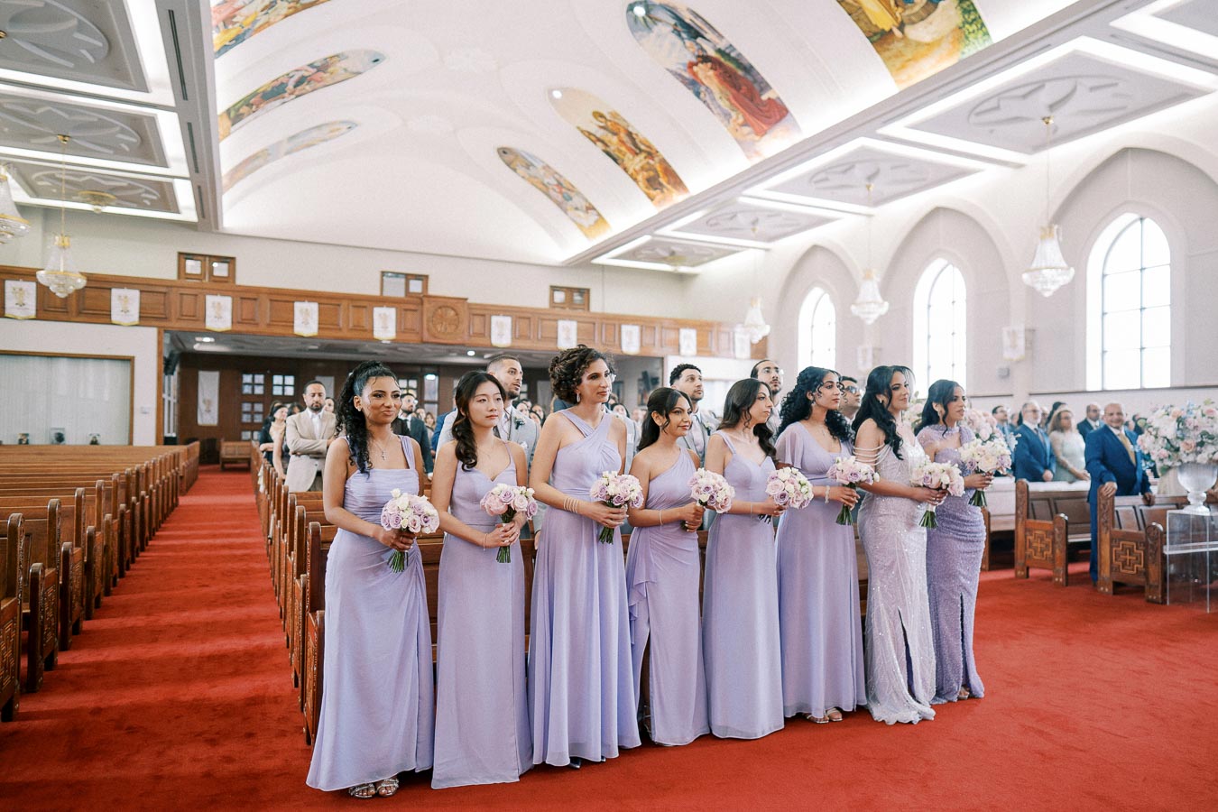 Bridesmaids in lavender dresses holding bouquets stand in a beautifully decorated church with a red carpet and ornate ceiling murals.