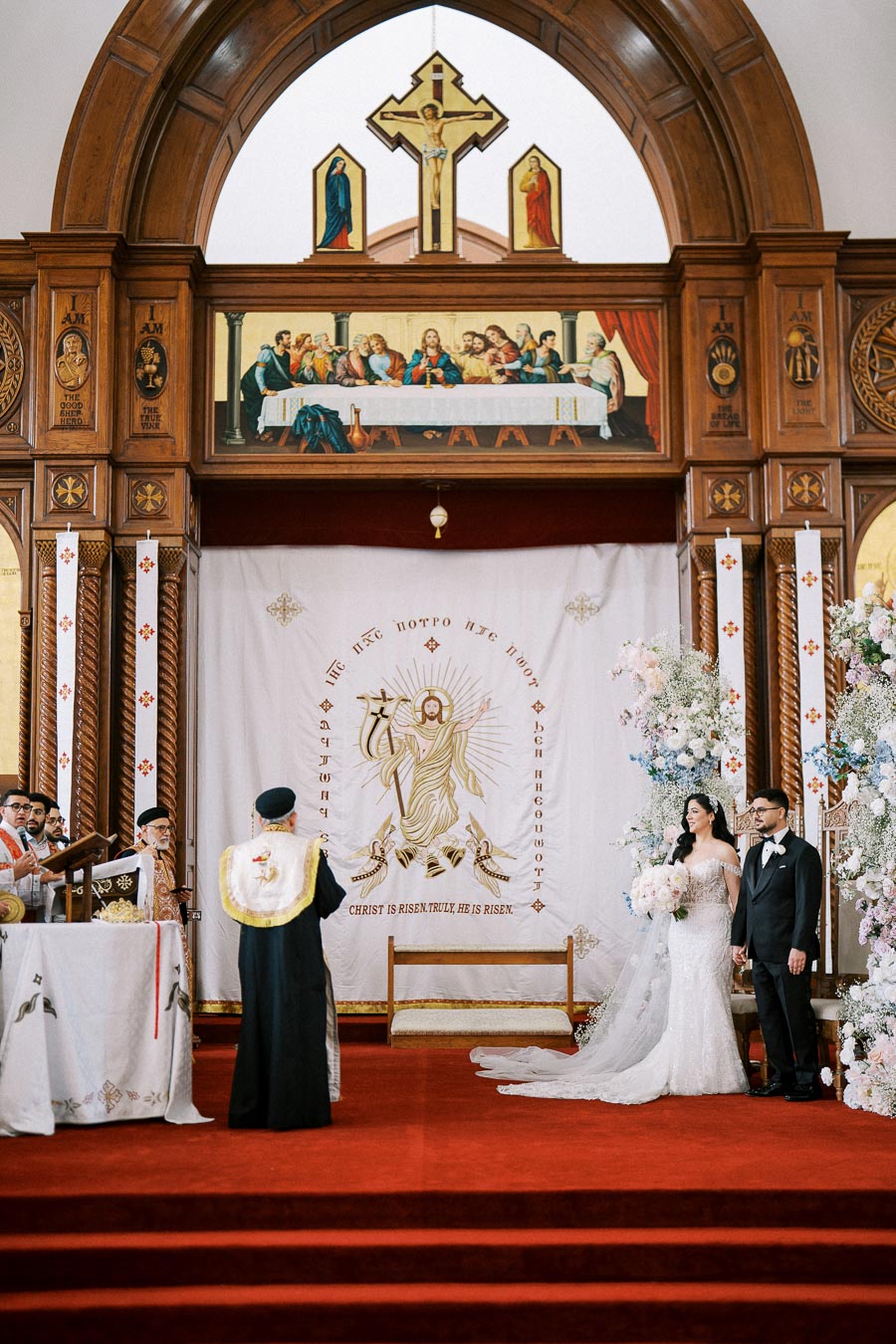 Wedding ceremony at ornate church altar with religious artwork and icons, featuring a priest officiating for a bride and groom standing near floral decorations.