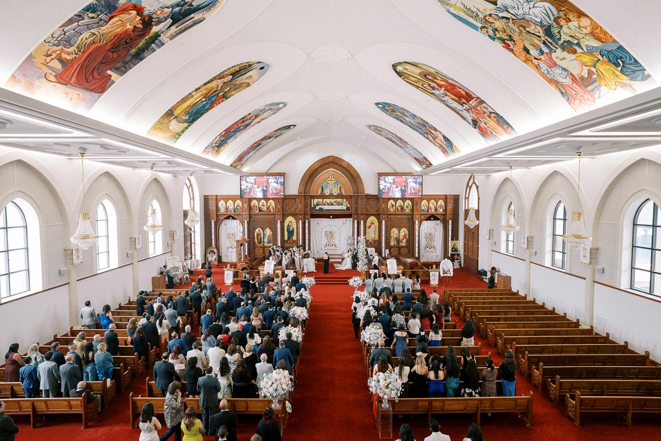 A large congregation gathers for a wedding ceremony in a beautifully adorned church with vibrant ceiling murals, arched windows, and decorated pews.